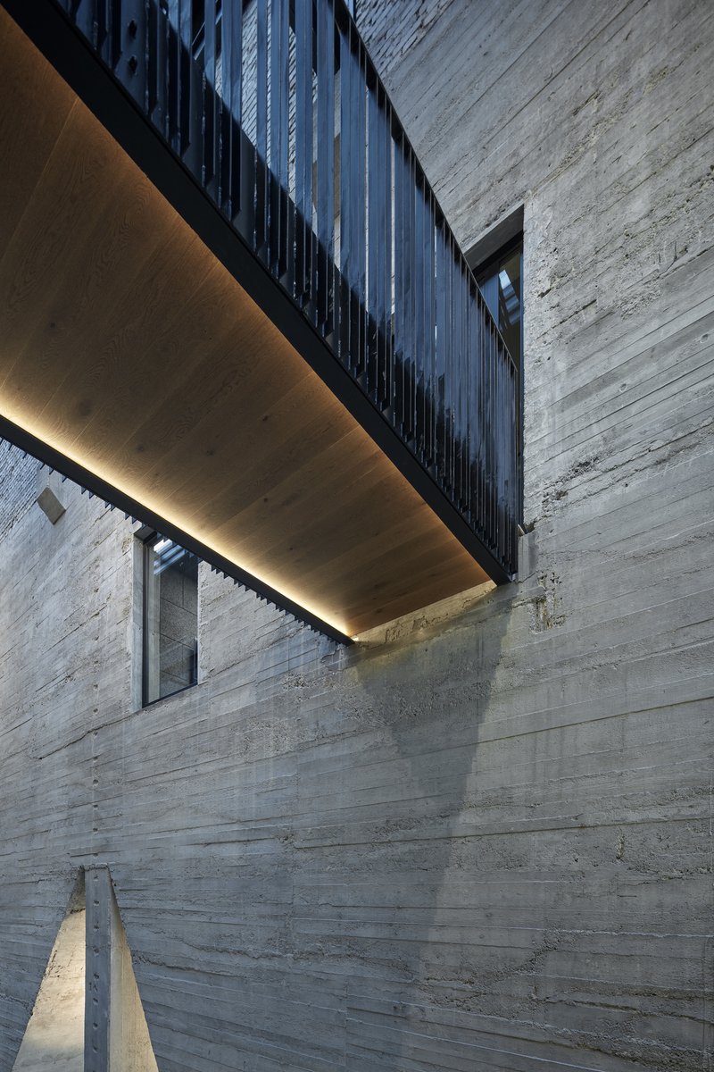 Cantilevered bridge with black metal cladding and warm linear lighting against boardformed concrete walls