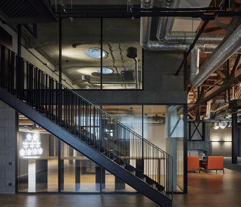 Black steel staircase with vertical balusters ascending past glass-enclosed meeting rooms under exposed structural ceiling