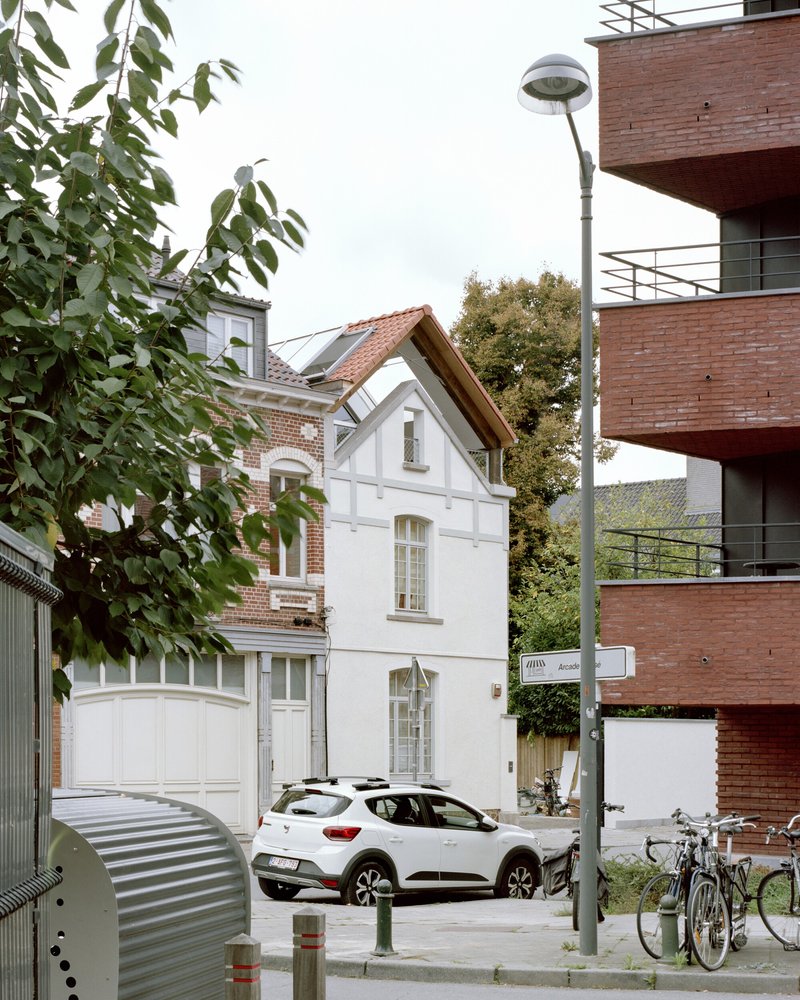 Street view of white rendered facade with gabled roof between brick neighbors and autumn trees