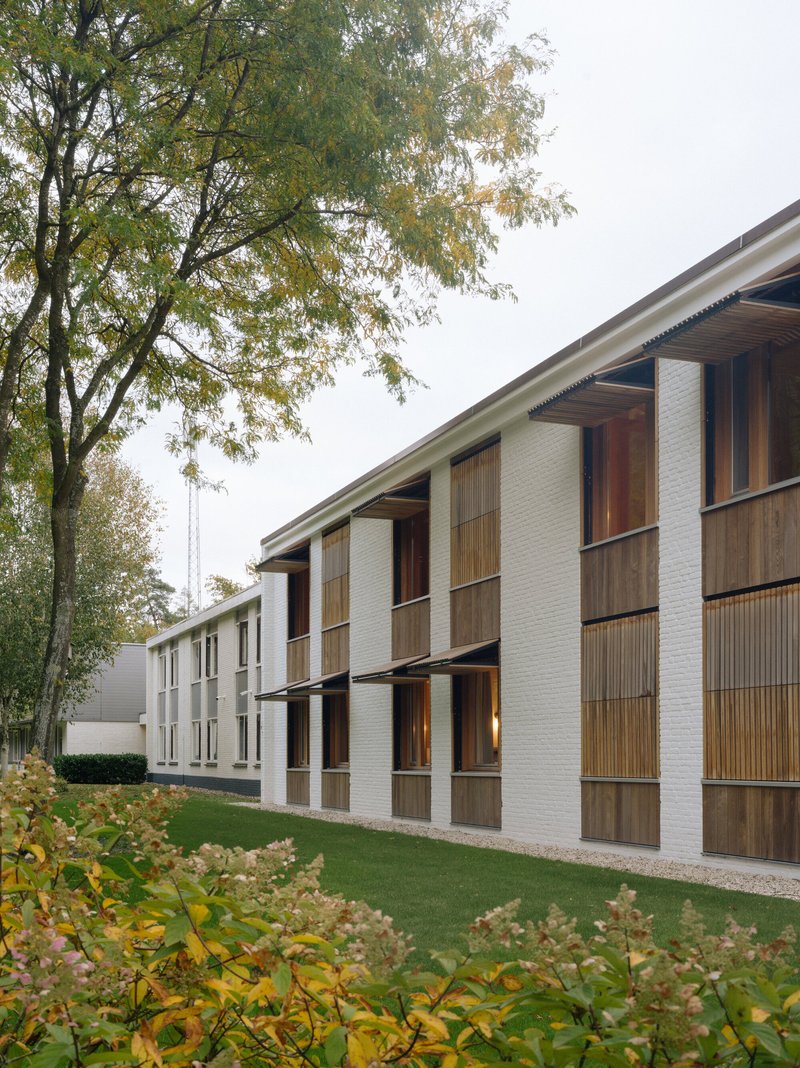 Two-story white brick facade with timber louvered balconies and vertical screens beneath autumn trees
