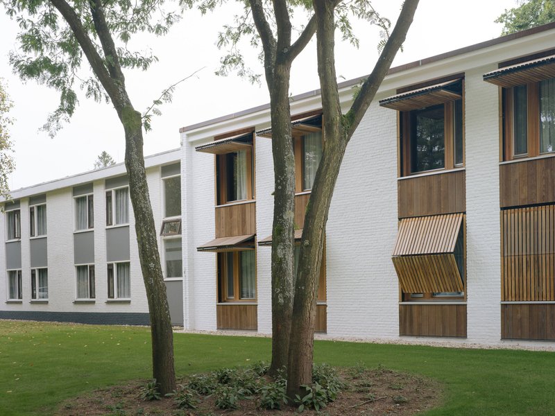 White brick facade with timber-clad bay windows and slatted screens framed by mature trees on a lawn