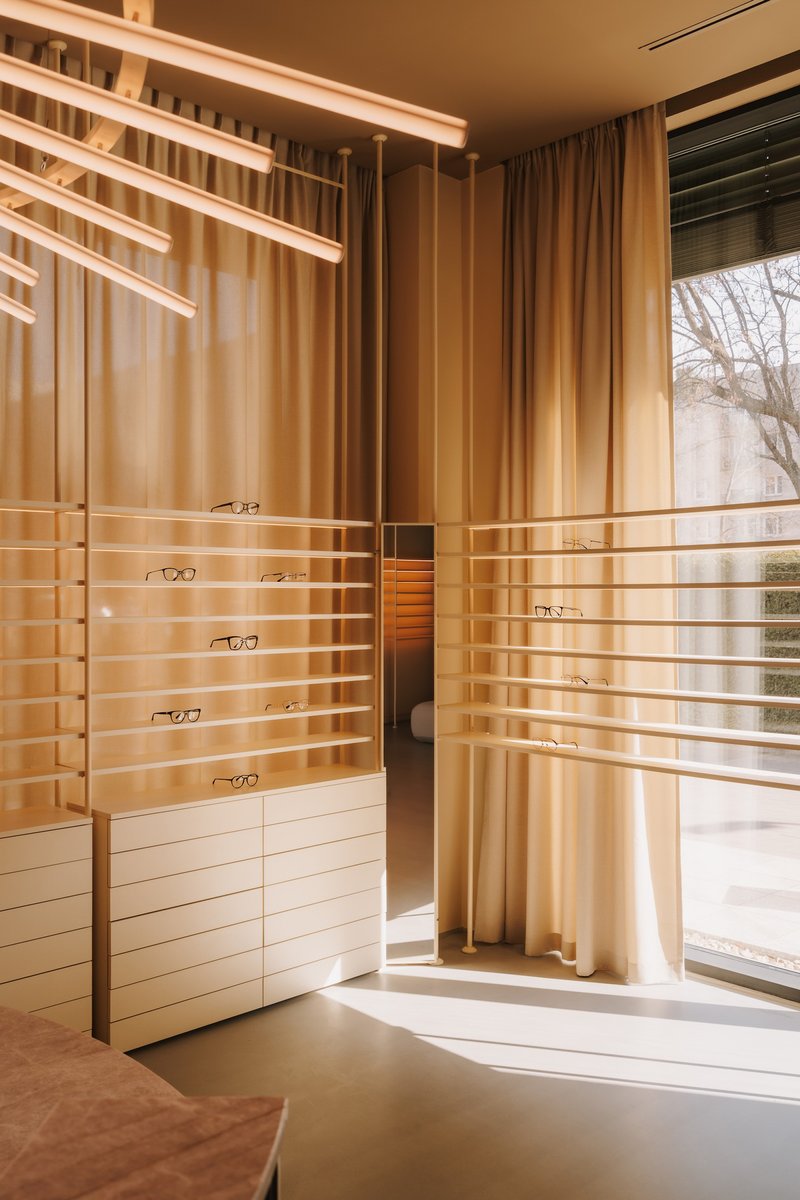 Corner display alcove with horizontal brass rods holding eyewear against floor-to-ceiling curtains and winter daylight