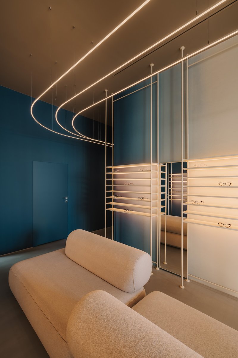 Curved suspended light fixtures above a cream sofa and horizontal metal shelving displaying eyewear