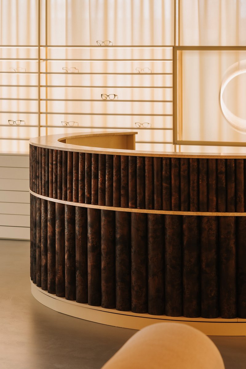 Curved reception desk with fluted dark wood panels against horizontal shelving holding eyeglasses