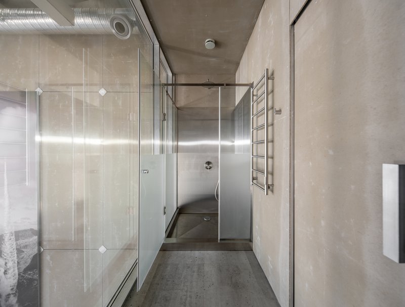 Corridor with glass shower enclosure, exposed concrete walls and ceiling, and industrial ducting overhead