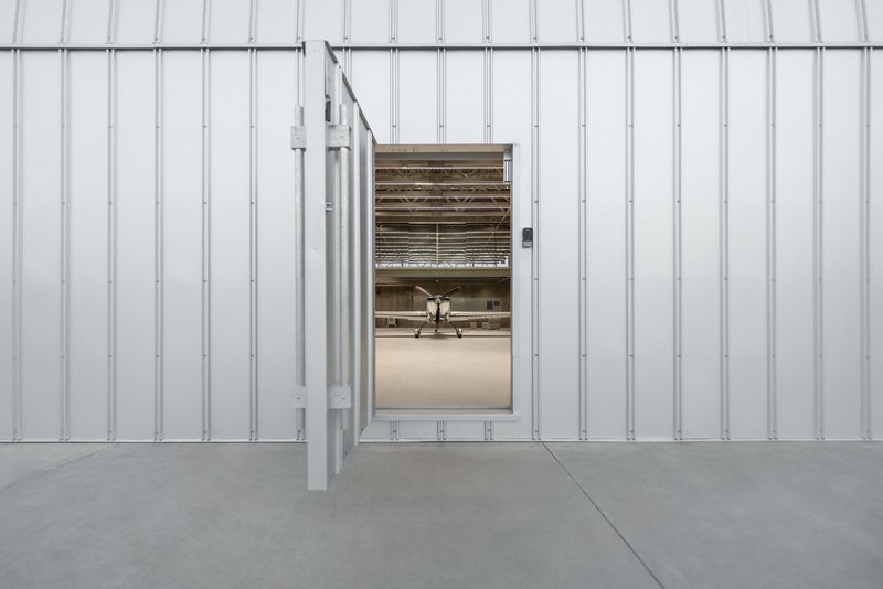 Corrugated metal cladding wall with open sliding door revealing aircraft inside the hangar bay