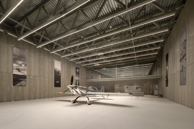 Long view down the hangar showing steel roof trusses and linear lighting above concrete walls