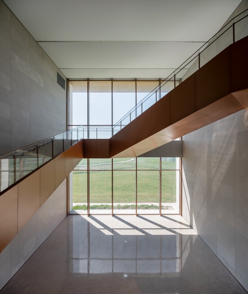 Double-height lobby with corten steel staircase and glazed curtain wall overlooking green lawn in daylight