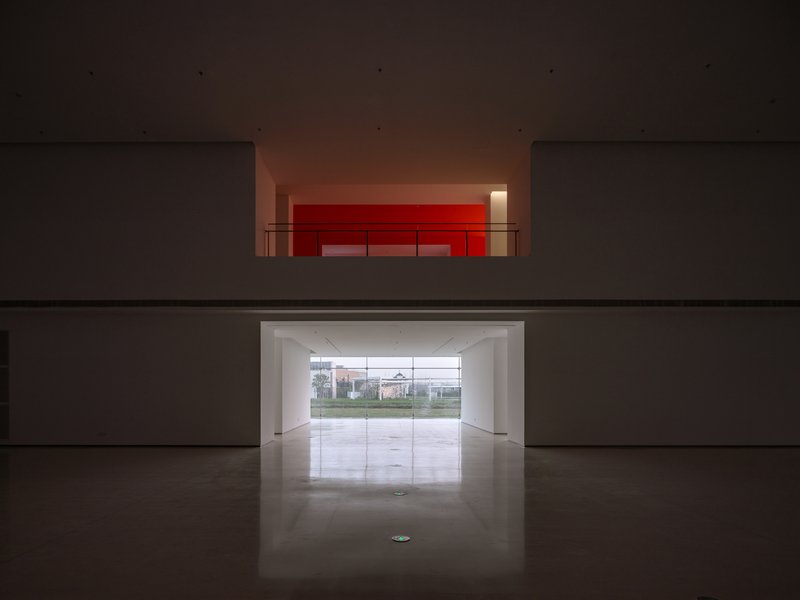 View through double-height void showing red-lit upper gallery and ground-level glazed entrance to landscape beyond