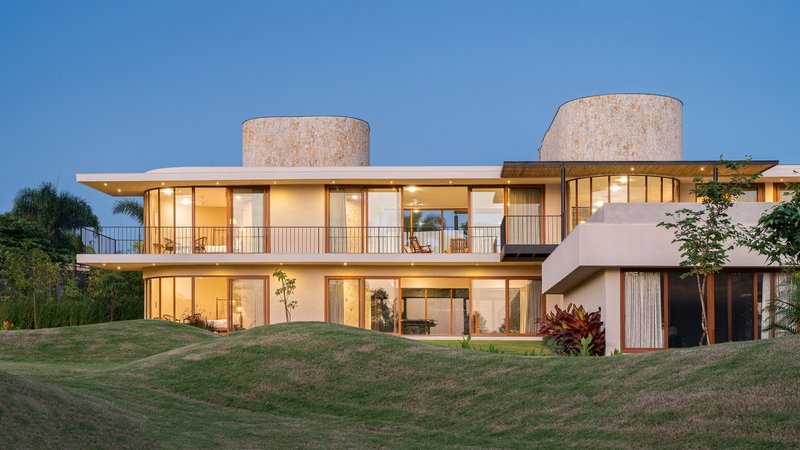 Two-story glazed facade with cylindrical stone towers and continuous balcony set on a sloping lawn at twilight