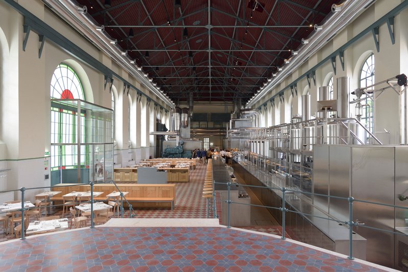Interior hall with stainless steel brewing equipment along one side and arched windows flooding the terracotta floor