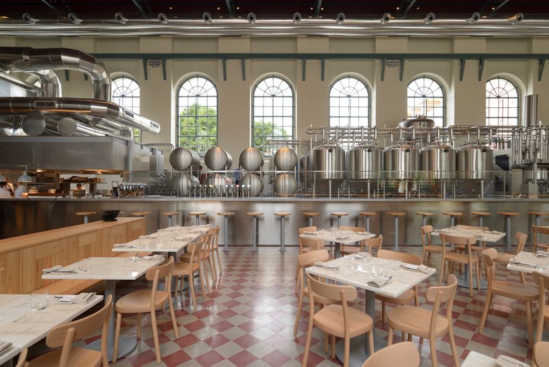 Dining area with timber tables and chairs on chequered flooring beside exposed stainless steel brewing tanks
