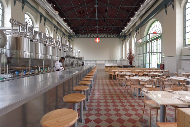 Long counter with circular stools facing stainless steel equipment under an exposed truss ceiling with red tiles