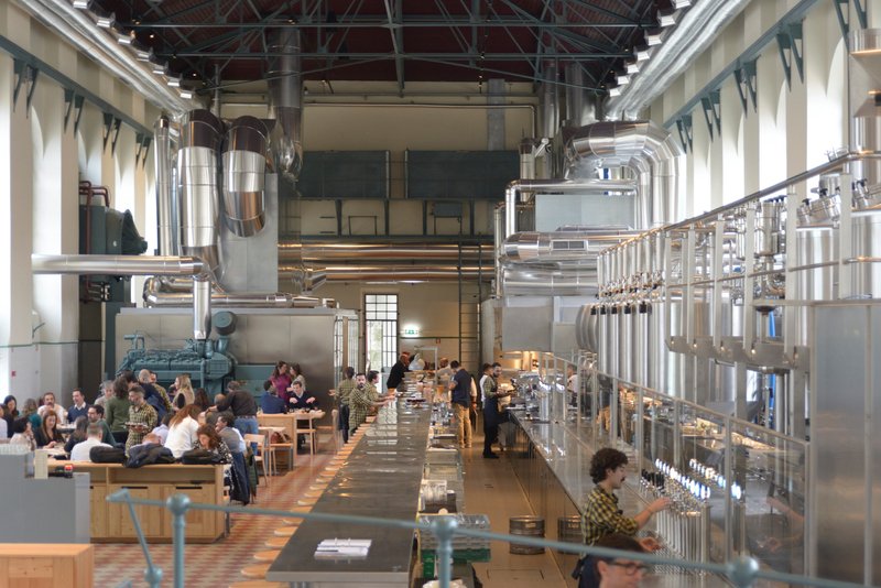Hall filled with diners at timber tables surrounded by elevated stainless steel tanks and industrial piping