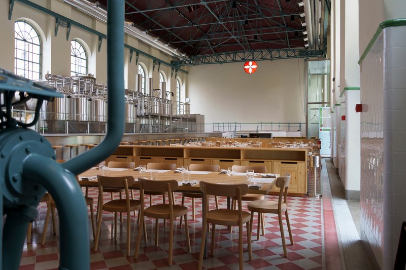 Round timber table with pink chairs set beside dark blue industrial pipework and brewing vessels
