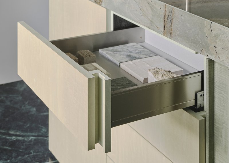 The image showcases a modern kitchen drawer, partially open to reveal an array of stone samples. The drawer features a light wood grain finish and is complement
