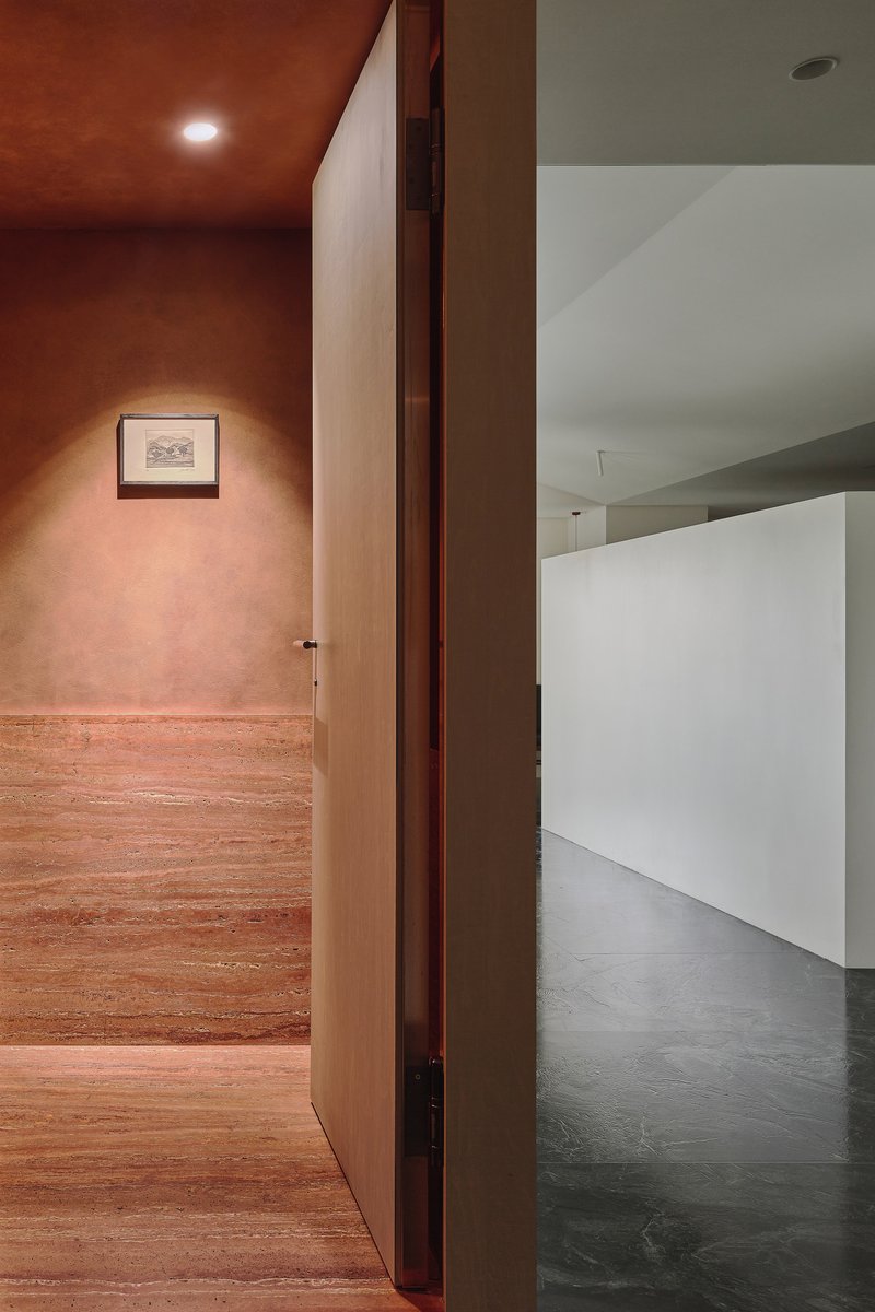 This architectural photograph showcases a doorway leading from a warm-toned, textured hallway into a minimalist, white-walled gallery space. The contrast betwee