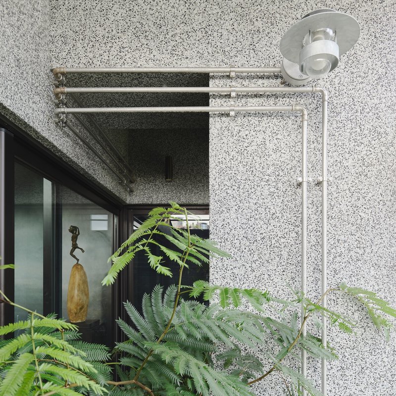 This architectural image showcases a modern building exterior with a textured terrazzo facade. A series of metal pipes and an outdoor light fixture are mounted