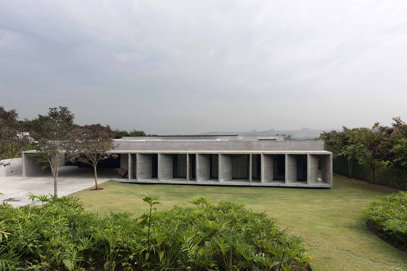 Long concrete pavilion with rhythmic columns set in lawn beneath trees and overcast sky