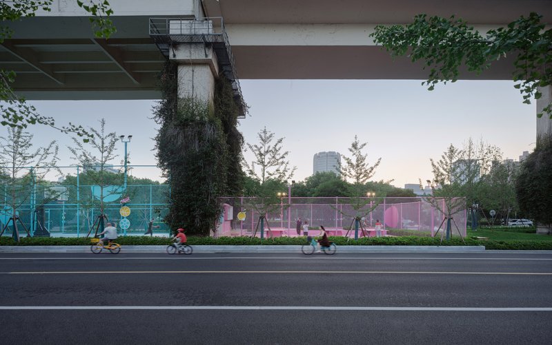 Street view of the pink mesh-fenced sports court beneath the elevated roadway with cyclists passing