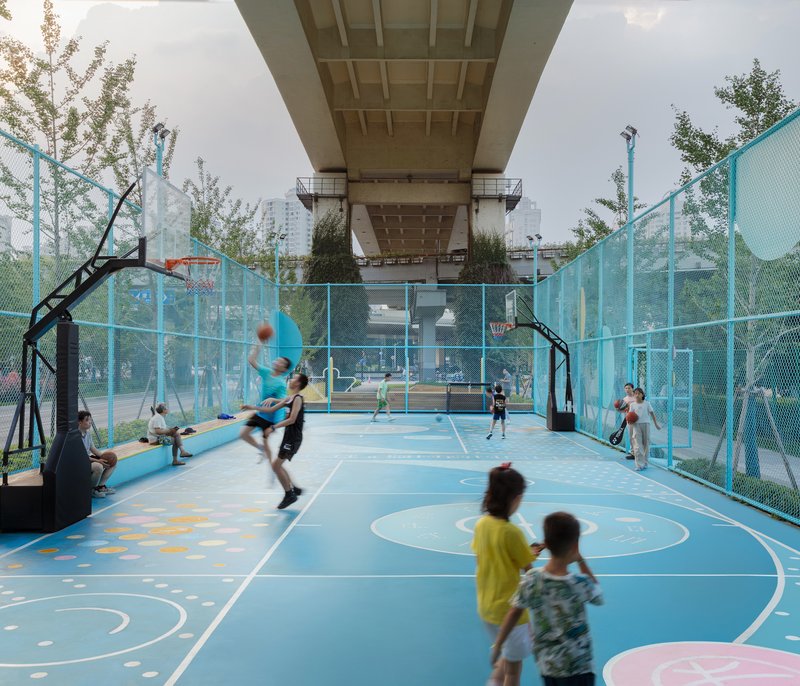 Blue basketball court with players in action beneath the concrete overpass structure on an overcast day