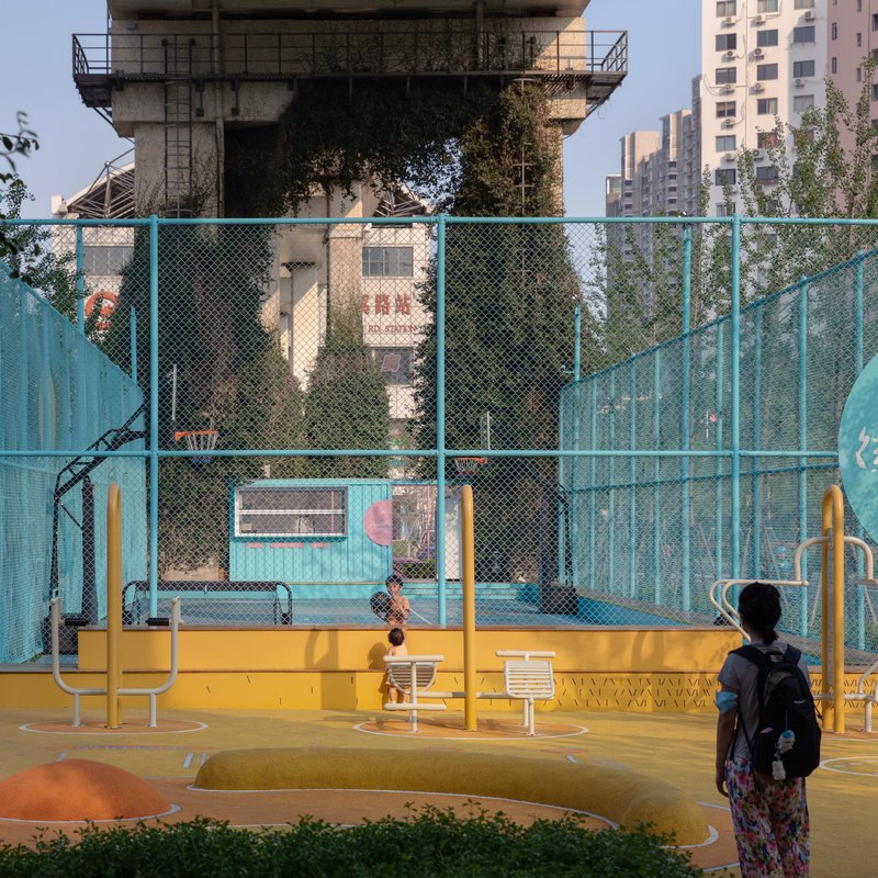 Outdoor basketball court with yellow base and turquoise mesh fencing beneath a concrete elevated structure