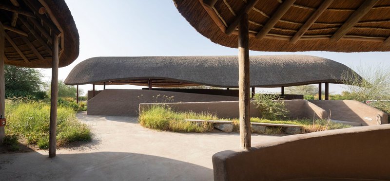 Courtyard view beneath circular thatched pavilions with curved earthen walls and planted beds in afternoon light