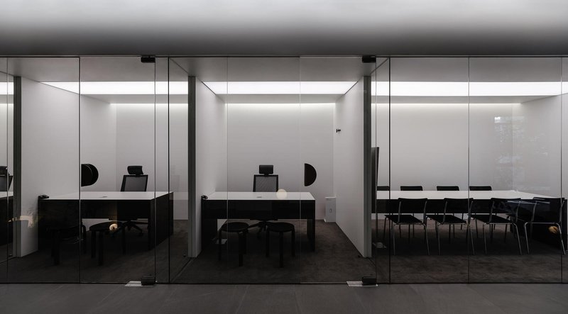 Glass-walled office spaces with black desks and chairs along a grey tiled corridor