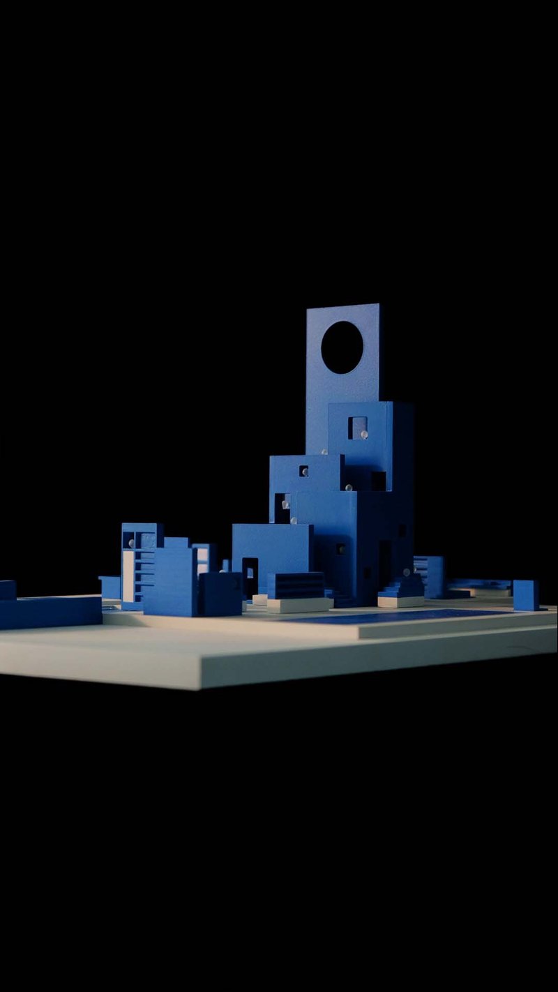 Physical model of a blue tower with circular portal and clustered volumes under dramatic side lighting