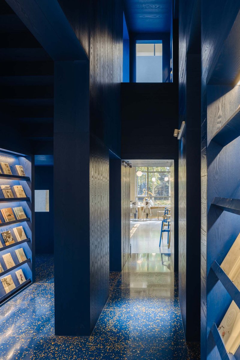Narrow corridor with blue terrazzo flooring, dark timber walls, and shelves leading to daylit space