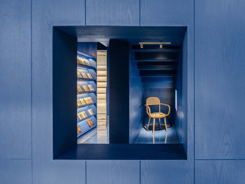 Framed view through blue wood paneling revealing illuminated shelves and a single bentwood chair