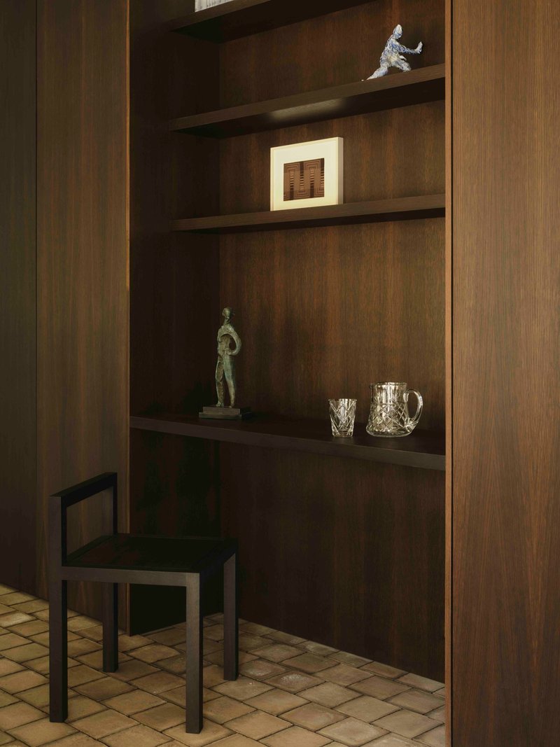 Dark timber shelving unit with display objects and small black chair against terracotta tile floor