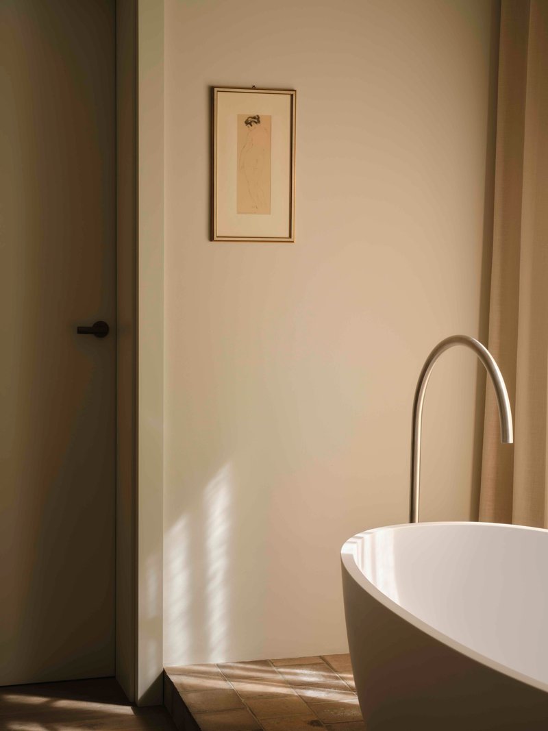 Freestanding white bathtub with arched floor-mounted faucet in soft afternoon light beside a framed drawing