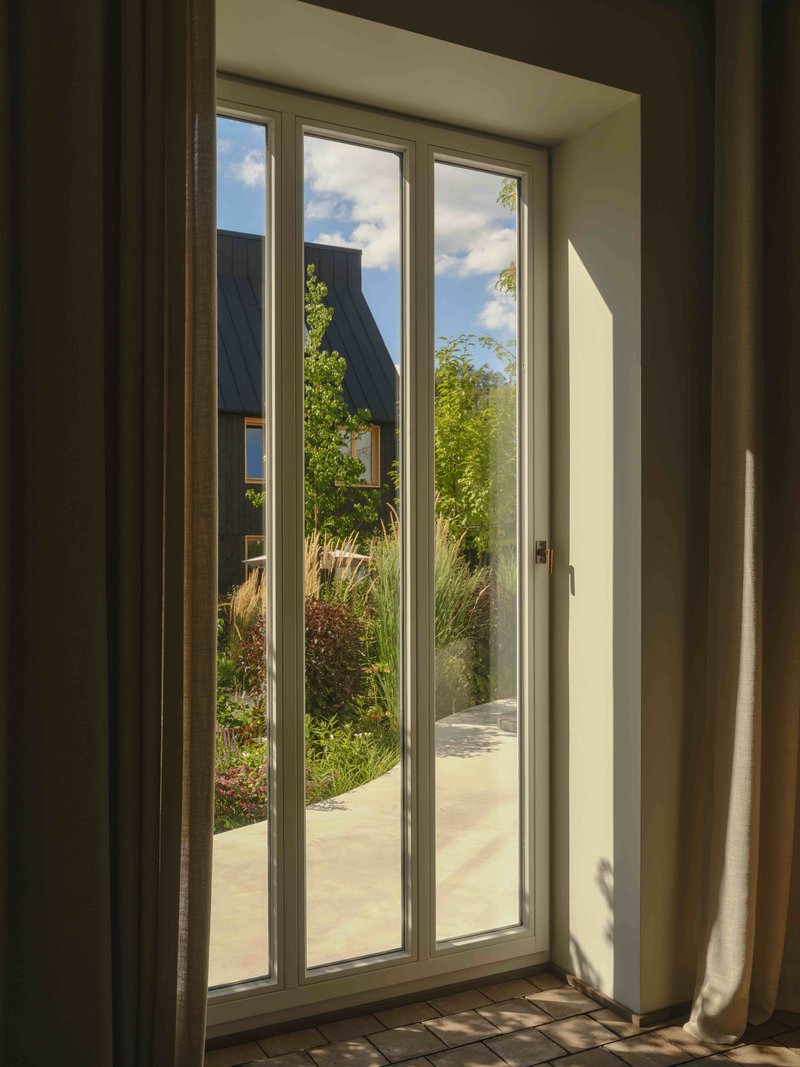 Triple casement window with white curtains framing a view of a courtyard garden and pitched roof beyond