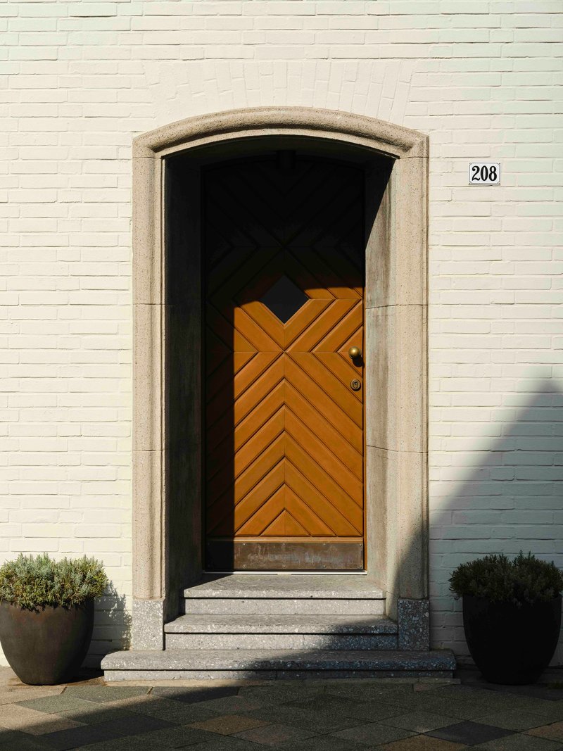 Arched doorway with chevron-patterned timber door set in white brick facade flanked by planters