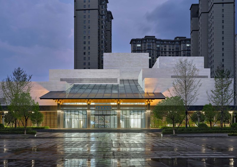 Glass entrance canopy illuminated at dusk with wet reflective paving in foreground and residential towers behind