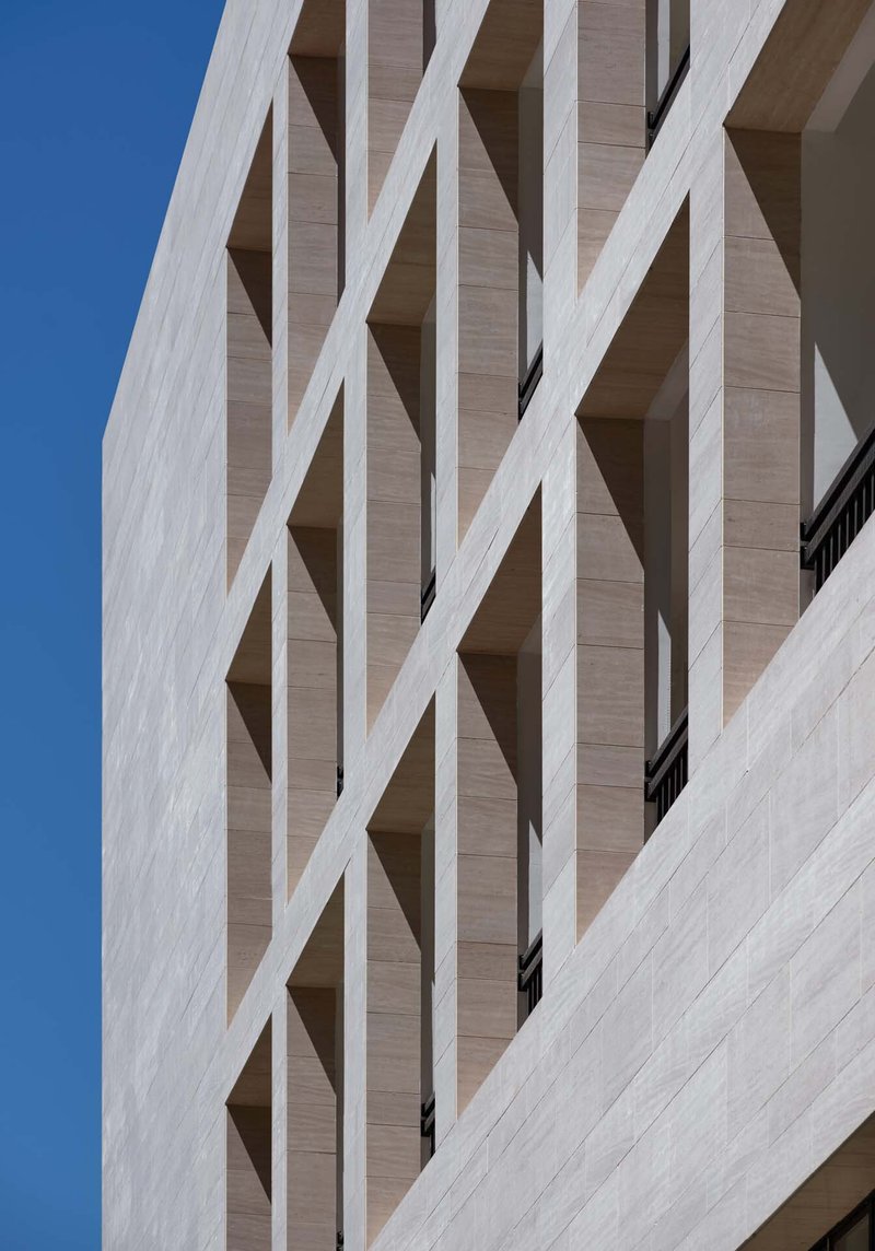 Detail of the travertine facade with diagonal white bracing elements forming an X-pattern against a clear blue sky