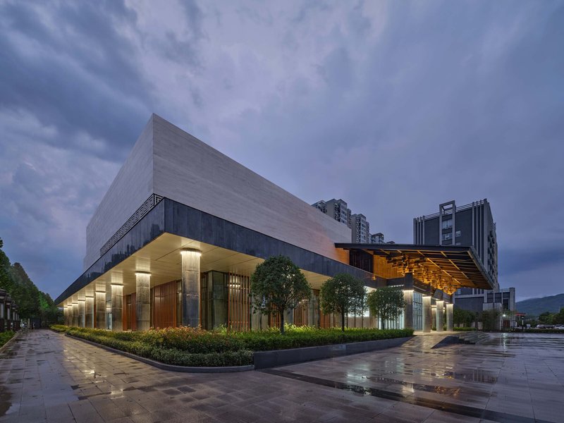 Corner view of the illuminated entrance colonnade with timber screens and planted beds at dusk after rain