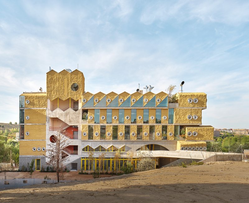Front facade with zigzag roofline, circular windows, and layered volumes of yellow brick and concrete in afternoon light
