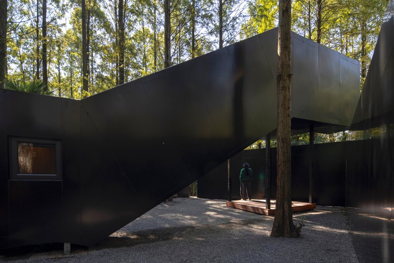 Covered walkway with undulating black metal walls and a figure standing beneath filtered afternoon light