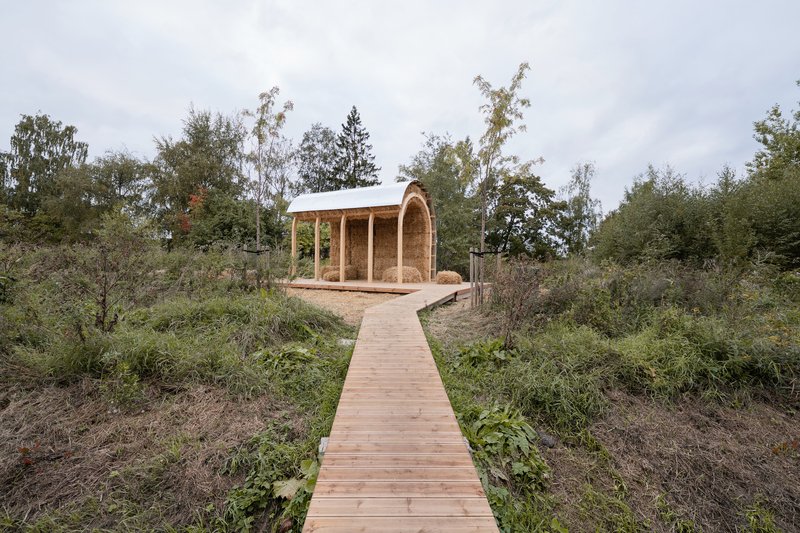 Timber pavilion with arched straw bale walls reached by a wooden boardwalk through planted landscape