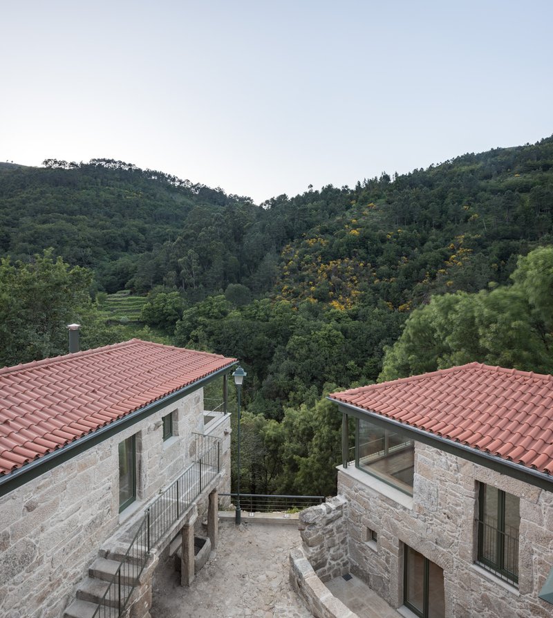 Courtyard between two stone volumes with terracotta roofs opening toward the valley beyond at dusk