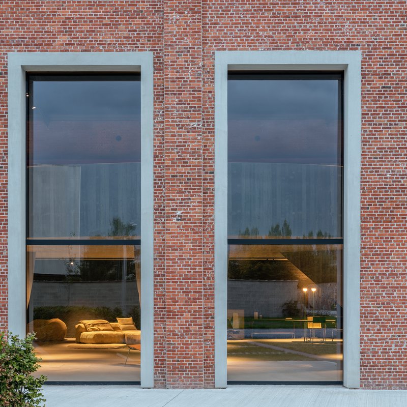 Two tall glazed openings framed in pale plaster reveal interior lounge furniture at dusk
