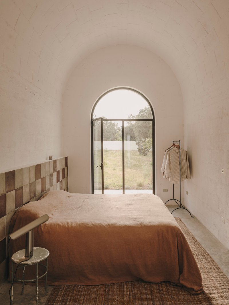 Bedroom with patchwork timber headboard and arched steel door framing view of olive grove