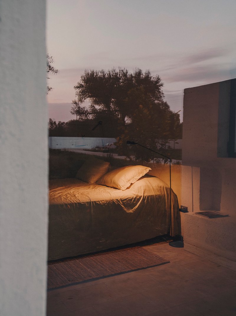 Bedroom with unmade linen bedding glimpsed through doorway at dusk with trees beyond glass wall