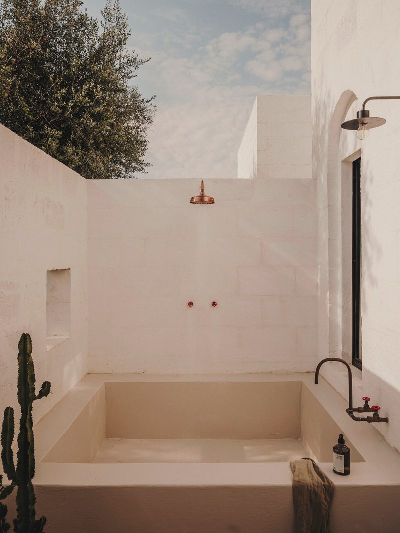 Outdoor bathing area with sunken tub, copper rain showerhead, and a cactus beside white plastered walls