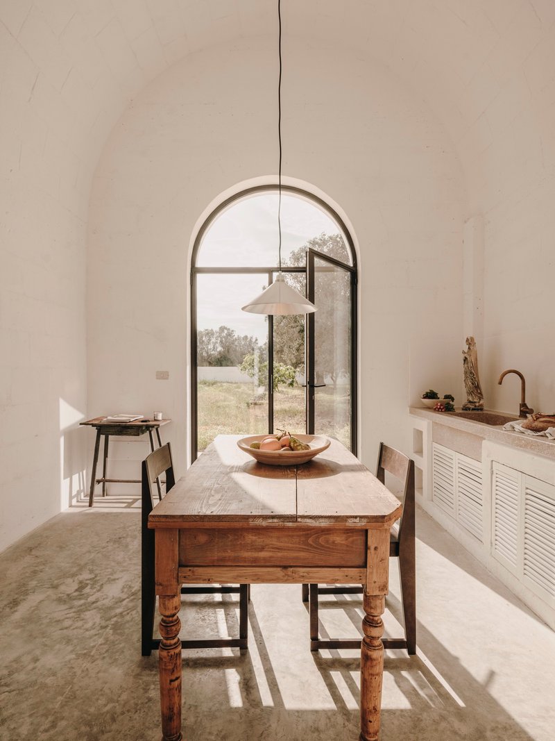 Dining area with wooden farmhouse table, pendant lamp, and arched glazed door overlooking an olive grove