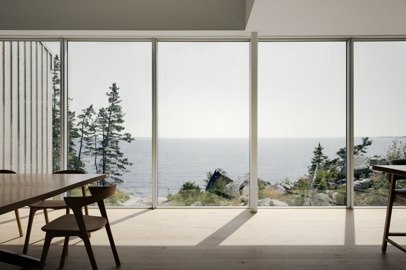 Floor-to-ceiling glass wall framing distant water and coastal pines with timber chair in sunlight