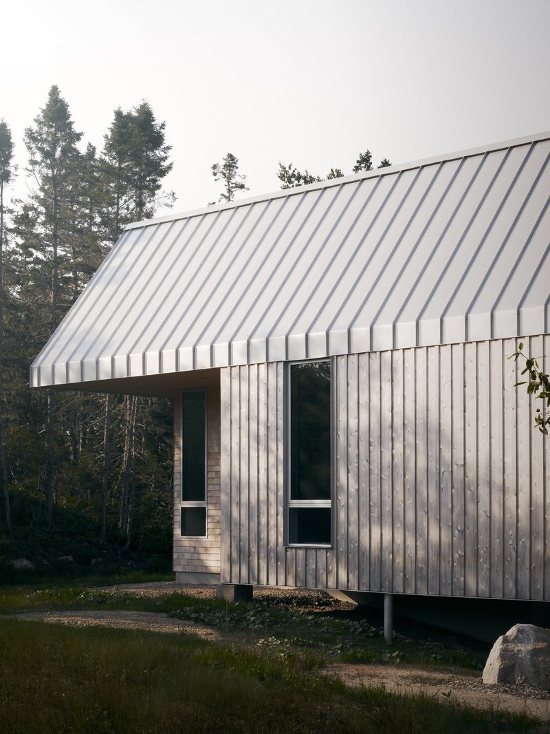 Gable-roofed volume with vertical timber siding elevated on concrete piers among evergreens