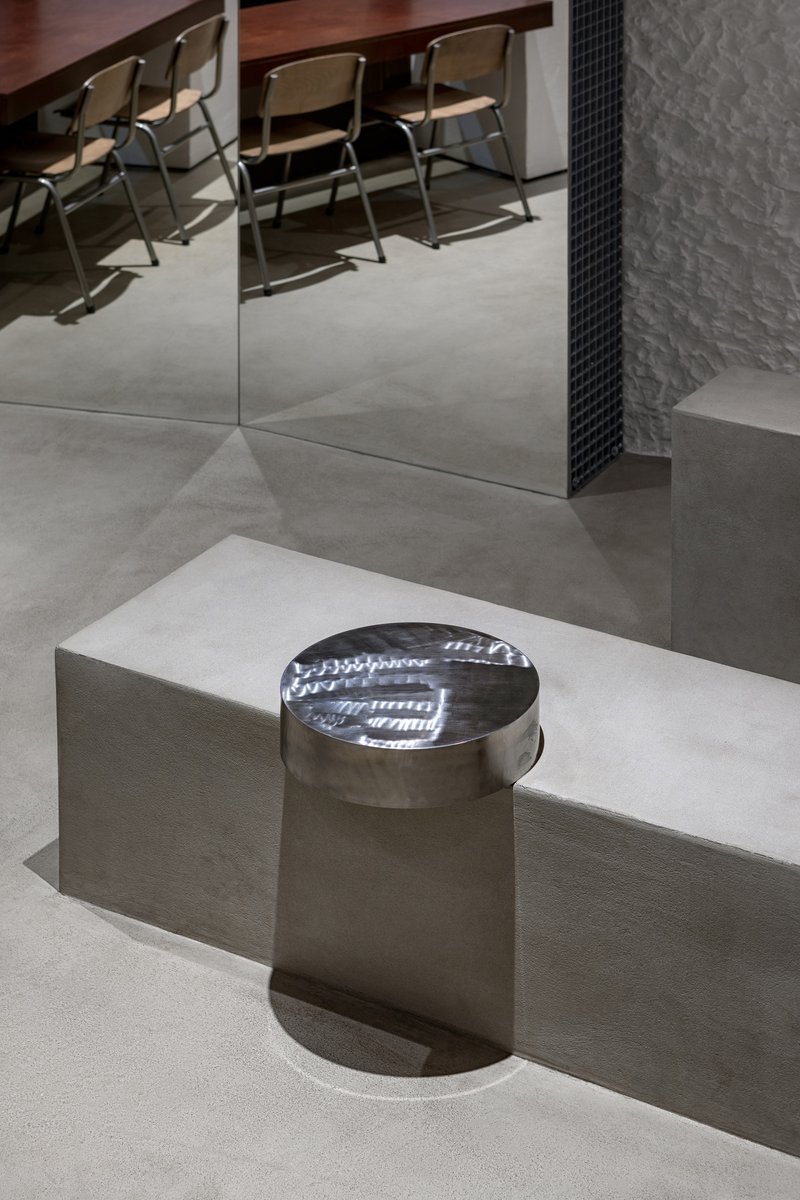 Polished metal cylindrical stool on a raised concrete platform beneath mirrored walls and folding chairs